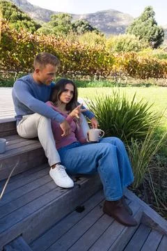 Diverse couple sitting on deck steps by vineyard in fall wear, woman pointing, Stock Photos