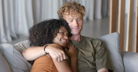Diverse couple sitting on gray sofa at home, man gesturing playfully, woman Stock Footage 338168383
