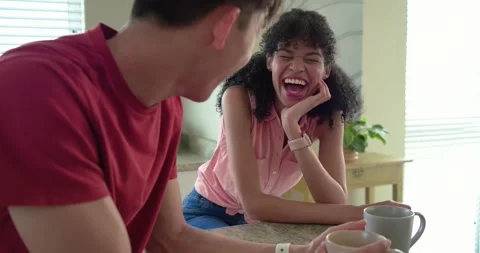 Diverse couple sitting at kitchen counter, man in red shirt telling joke woman Stock Footage 340773362