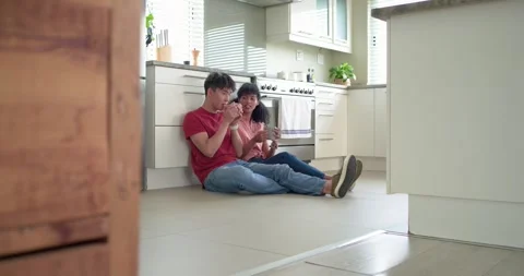 Diverse couple sitting on kitchen floor with mugs, man showing smartphone to Stock Footage 340069519