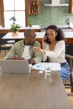 Diverse couple sitting at kitchen table, working with laptop, pie chart papers, Stock Photos