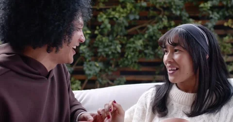 Diverse couple sitting on light couch at leafy patio, woman teasing, touching Stock-Footage 345607898