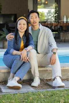 Diverse couple sitting on pool coping at backyard pool, wearing white sneakers Foto stock