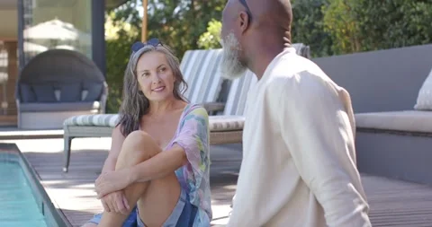 Diverse couple sitting at pool deck, man turning head starting conversation to Stock Footage 337933153