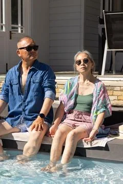 Diverse couple sitting on pool ledge in backyard, feet in water, wearing Foto stock