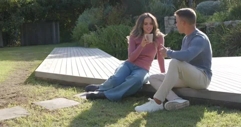 Diverse couple sitting on raised deck in backyard, man offering mug, woman Vídeo Stock 344255099