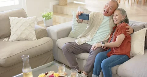 Diverse couple sitting on sectional sofa, man picking blue phone, taking selfie Vídeos de archivo 341090785