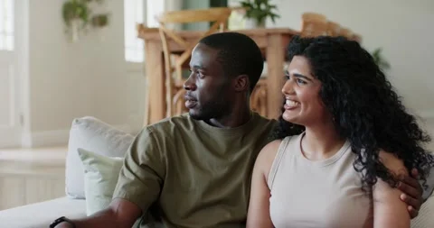 Diverse couple sitting on sofa at home, reacting to off-camera motion, man Stock Footage 340773922