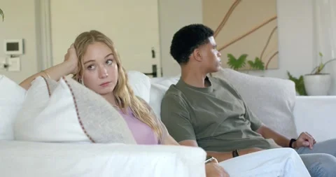 Diverse couple sitting on sofa at home after disagreement, woman glancing, man Stock Footage 341242366