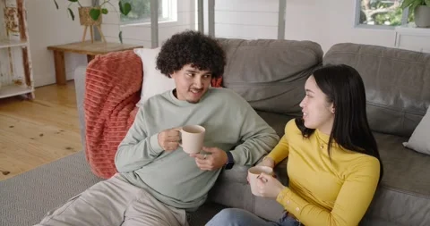 Diverse couple sitting on sofa with mugs, after eye contact man raising mug, Stock Footage 345090456