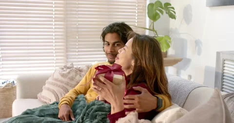 Diverse couple sitting on sofa, pulling gift from box with red ribbon to Stock Footage 340610963