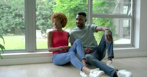 Diverse couple sitting by window at home, woman in red-top pointing at yard, man Stock Footage 338326018