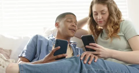 Diverse couple on sofa scrolling phones, man shifting gaze toward partner Stock Footage 344104831