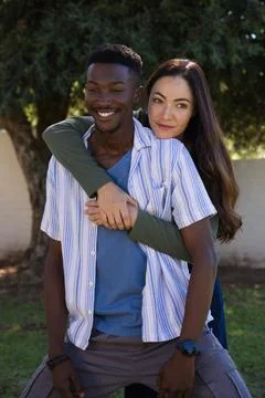 Diverse couple standing in backyard under large tree, woman hugging man, Stock Photos