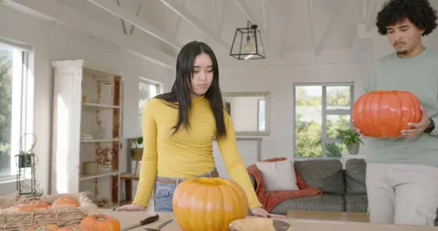 Diverse couple at table, man entering carrying pumpkin, scooping seeds with Vídeos de archivo 345090404