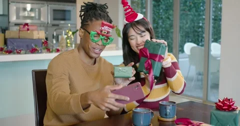 Diverse couple at table, man lifting phone, woman with mug showing wrapped gifts Stock Footage 342031776