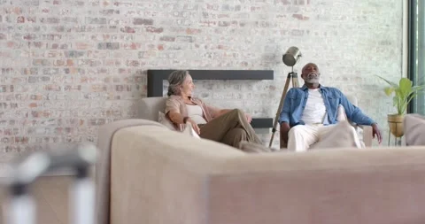 Diverse couple talking on sofa at home following suitcase handle signaling Vidéo 335071124