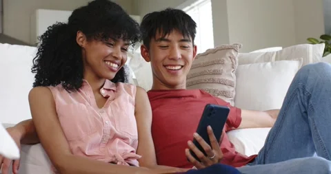 Diverse couple tapping and scrolling phone, laughing on cream-colored sofa in Stock Footage 341851837