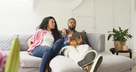 Diverse couple tapping tablet on couch as woman entering with phone, exchanging 스톡 동영상 317065353
