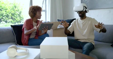 Diverse couple unpacking white VR headset at home on gray sofa, man testing, Stock Footage 344331175
