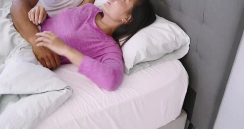 Diverse couple waking under white bedding, deactivating phone alarm, exchanging Stock Footage 336844278
