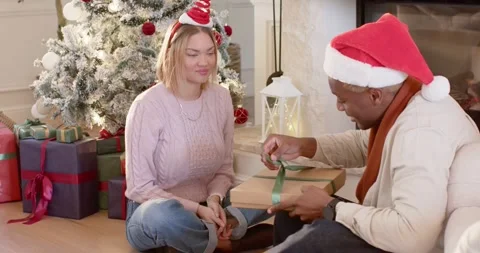 Diverse couple by Xmas tree during gift exchange, man in Santa hat untying green Stock Footage 343469410