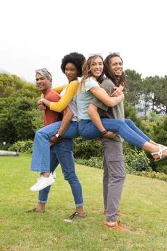 Diverse couples posing on grassy lawn in park, carrying partners while showing Stock Photos
