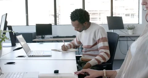 Diverse coworkers man writing in notepad at office desk by laptop smiling at Video stock 345589360