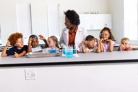 Diverse elementary kids with instructor in lab coat watching beakers at lab Foto stock