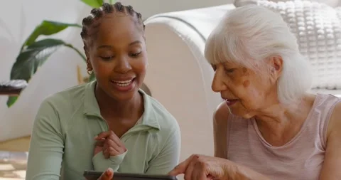 Diverse female aide showing tablet on sofa while senior woman pointing at Stock Footage 342494736