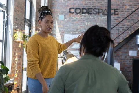 Diverse female colleagues presenting code with marker on glass board at cowor Stock Photos