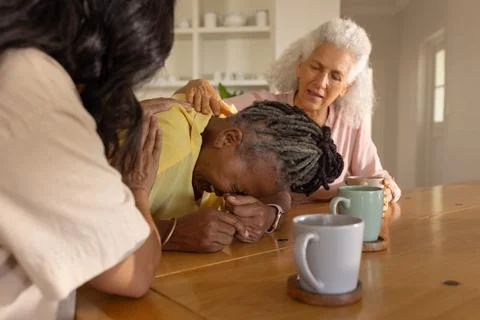 Diverse female friends consoling senior woman leaning over wooden table in Stock Photos