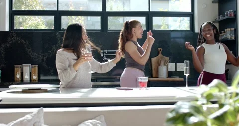 Diverse female friends dancing at kitchen island after phone starting music, Stock Footage 344340703
