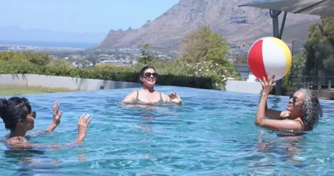 Diverse female friends in infinity pool tossing red beach ball, right woman Stock Footage 344331362