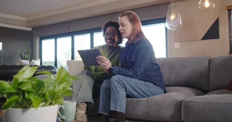 Diverse female partners scrolling tablet, reacting to content on grey sofa, Stock Footage 342031236