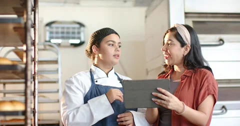 Diverse female team in bakery checking tablet, lead showing plan, chef in coat Stock Footage 341944666