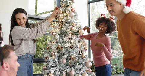 Diverse friends placing gift, hanging baubles, adjusting lights and admiring Stock Footage 338325622