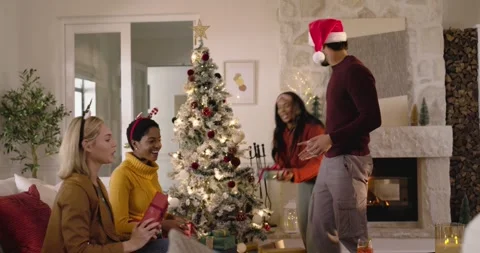 Diverse friends sitting by Christmas tree exchanging gifts while Santa hat man Stock Footage 344459949