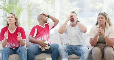 Diverse group on sofa two wearing red, with ball, watching match, reacting to Video stock 337625285