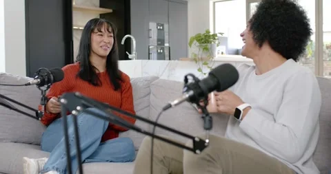 Diverse hosts podcasting on gray couch mics, man checking watch sparking smiles, Stock Footage 345607729