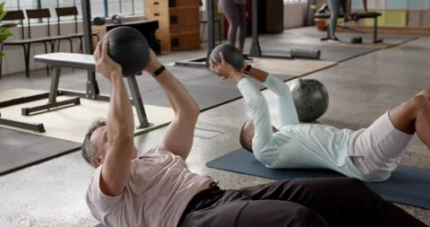Diverse men lying supine with black med balls, performing sit-up-to-press reps Stock Footage 337255453