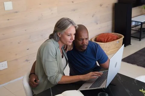 Diverse pair leaning over silver laptop at dining table, senior woman guiding Fotos Stock