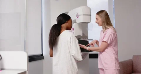 Diverse patient placing hand on mammogram; pink-scrub tech gesturing with tablet Stock Footage 337400178