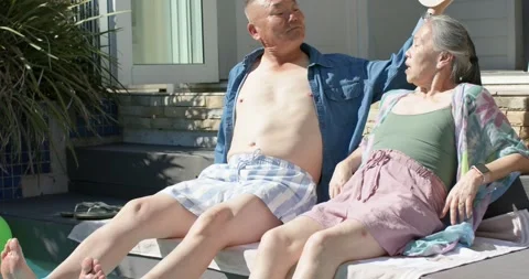Diverse senior couple sitting pool ledge, feet in water, man reaching, placing Stock Footage 335071146