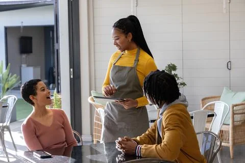 Diverse server and diners chatting at cafe, wearing mustard top, holding tablet Stock Photos