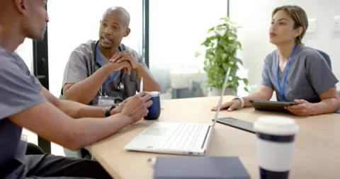 Diverse staff in scrubs at table discussing care prompted by laptop using tablet Stock Footage 345123811
