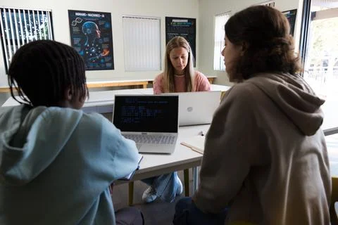 Diverse students collaborating at table in classroom, using laptops with code Photos