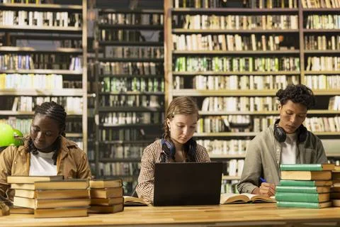 Diverse students learning at library table with textbooks and laptop Stock Photos