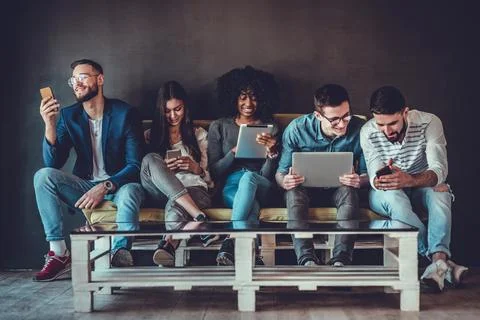 Diverse students using gadgets, sit on sofa in line on grey wall background i Stock Photos