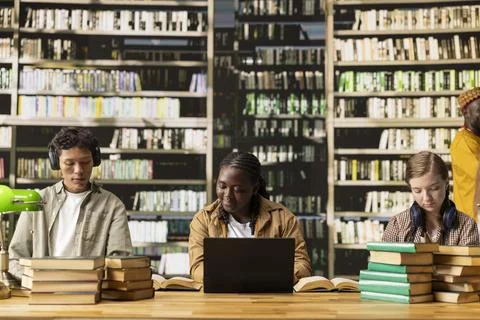 Diverse students working at shared library table with laptop and open books Stock Photos